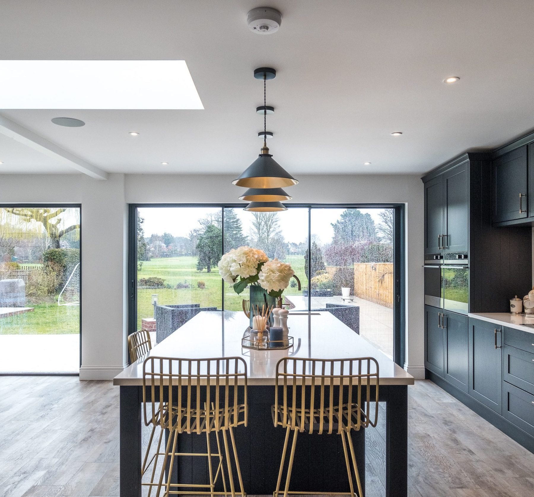 An internal view looking down the length of the kitchen island with feature light pendants above, and large glass sliding patio doors into the patio with the golf course in the background.