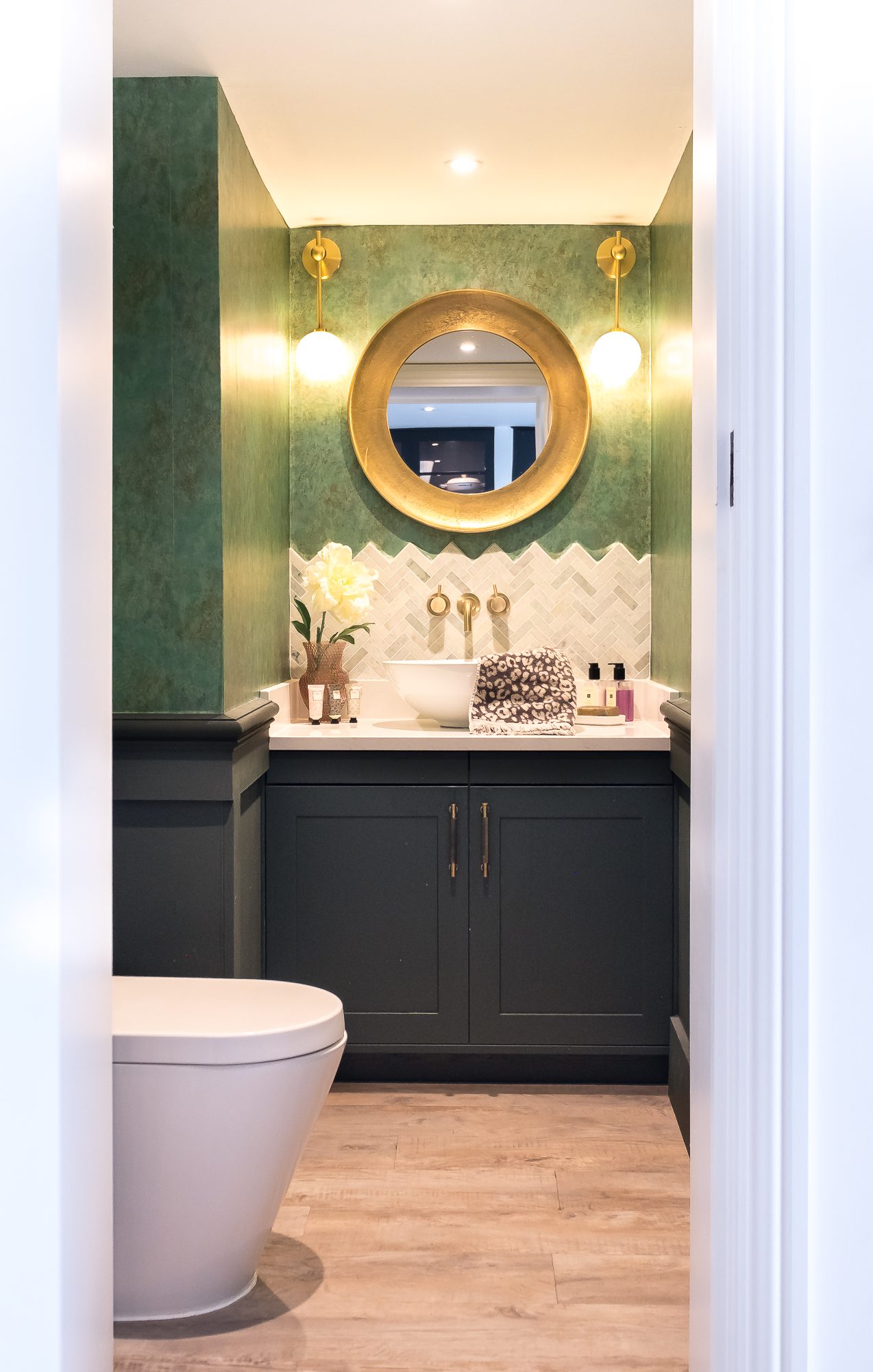 An internal view of the ground floor cloak room, with dark coloured walls and an opulent gold mirror and brushed brass light fittings and sanitaryware at Topsham Road.
